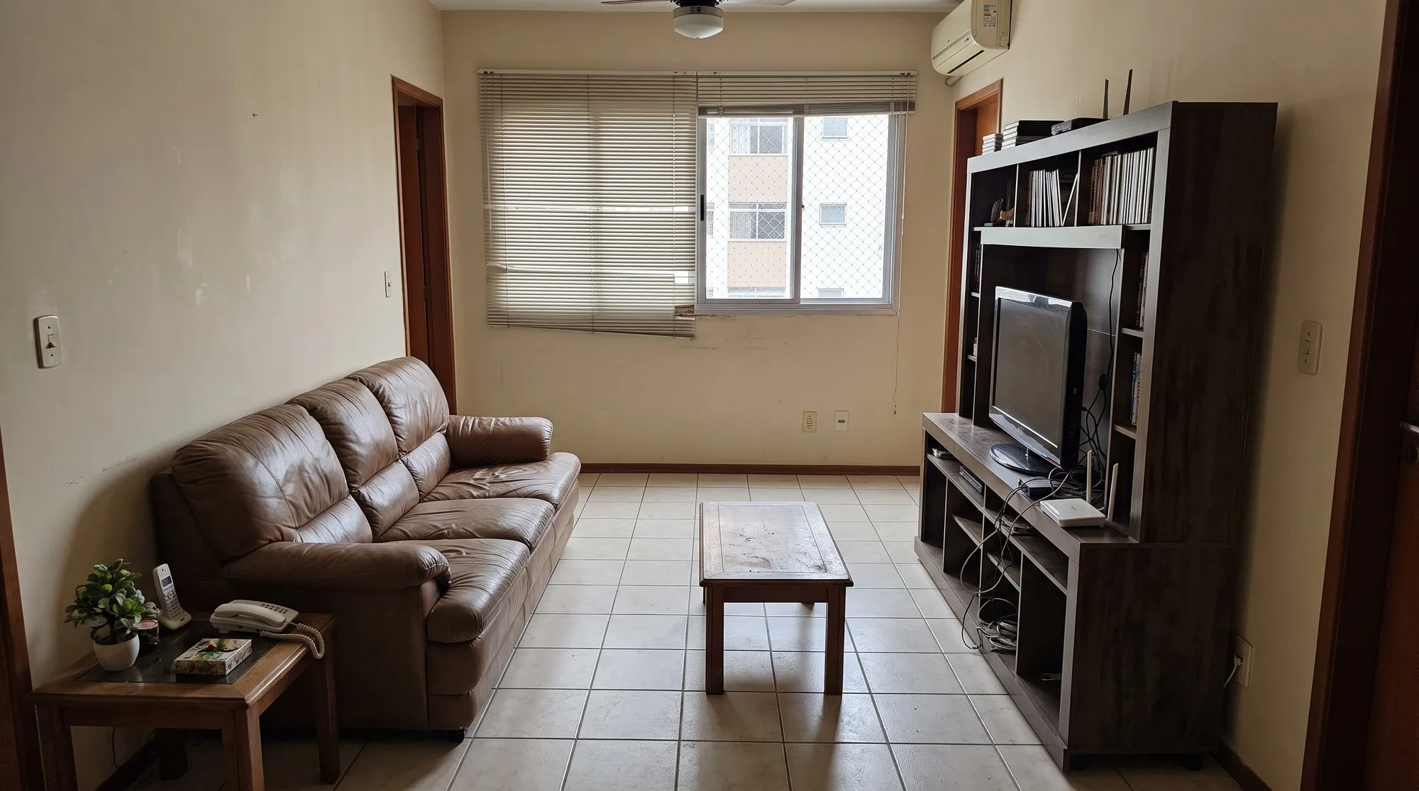 Sala de apartamento com sofá de couro marrom, rack escuro com TV e paredes bege