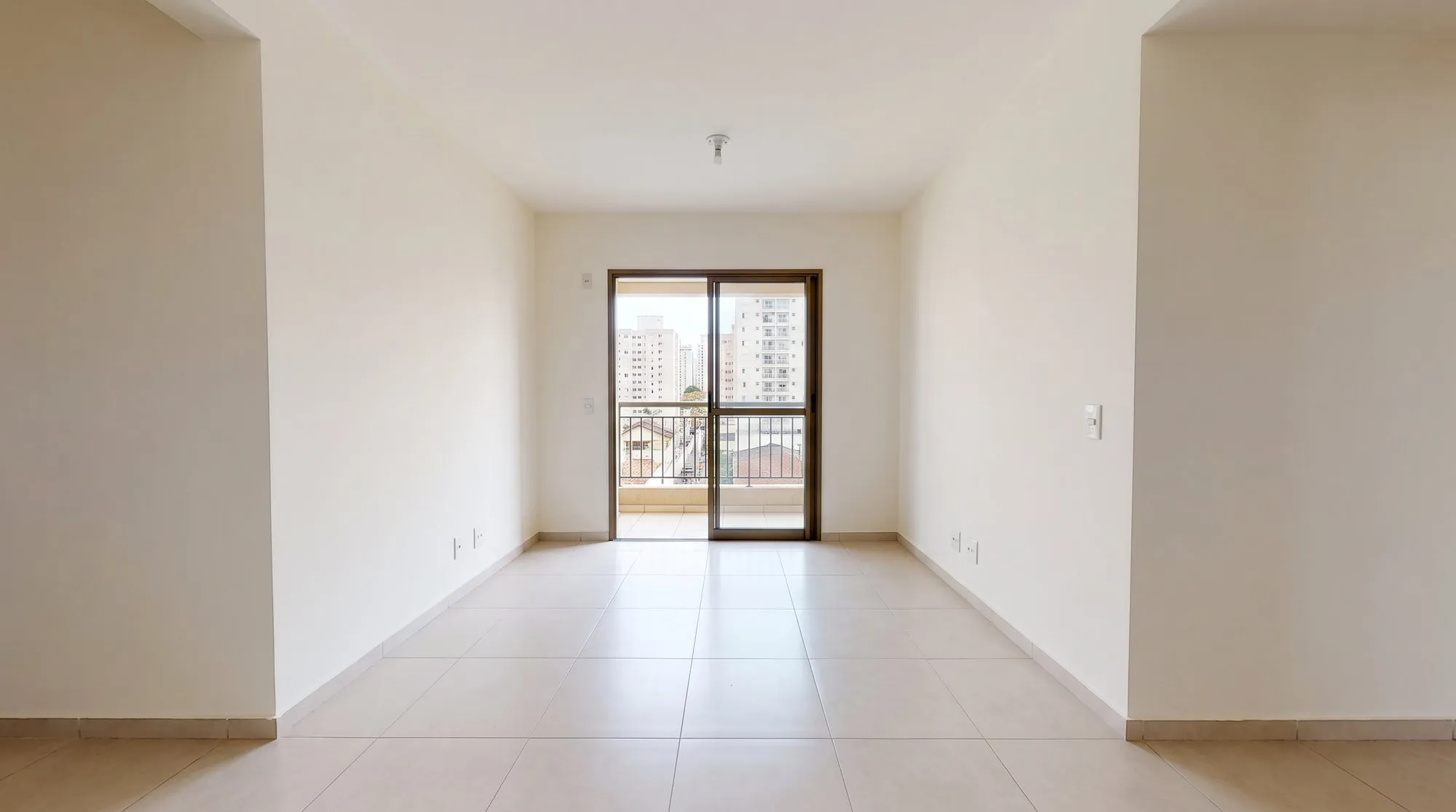 Sala vazia de apartamento, fotografada da porta, com piso claro e a varanda ao fundo